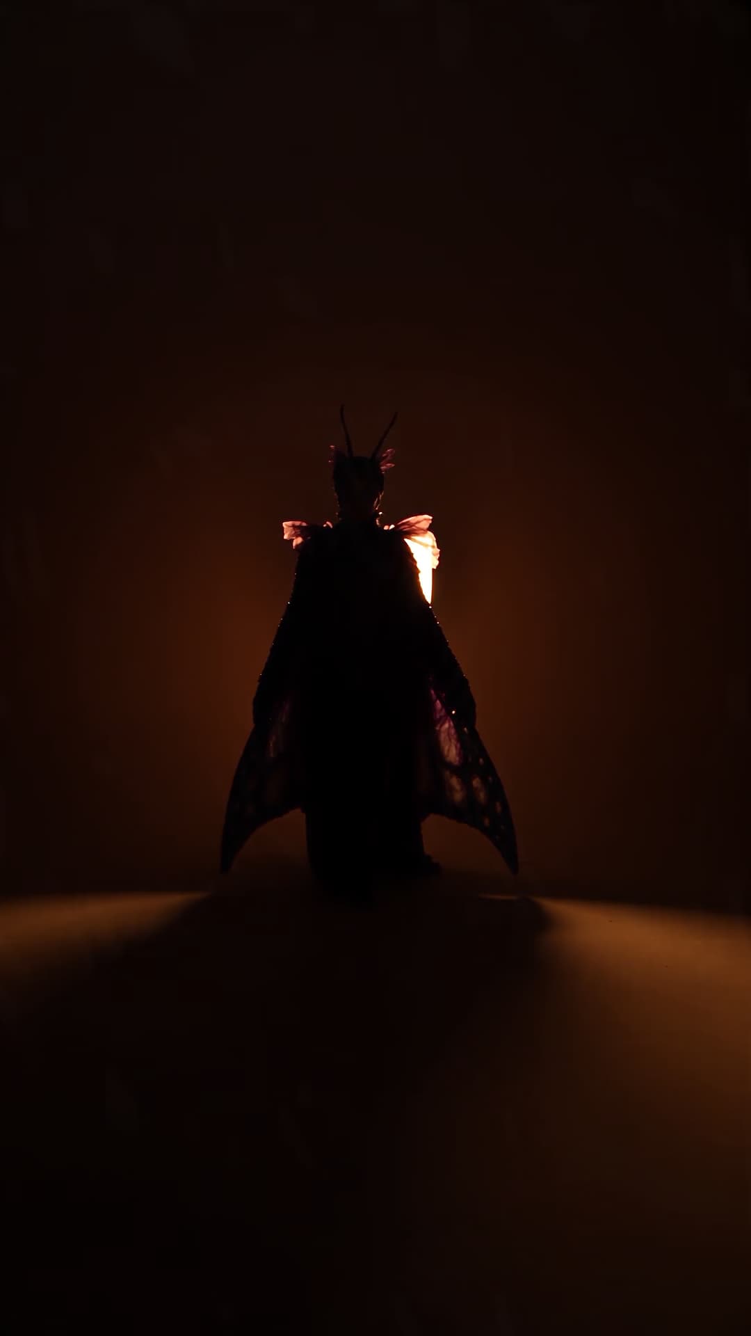 Creator in an original celestial moth costume opening orange and violet wings in warm spotlight.