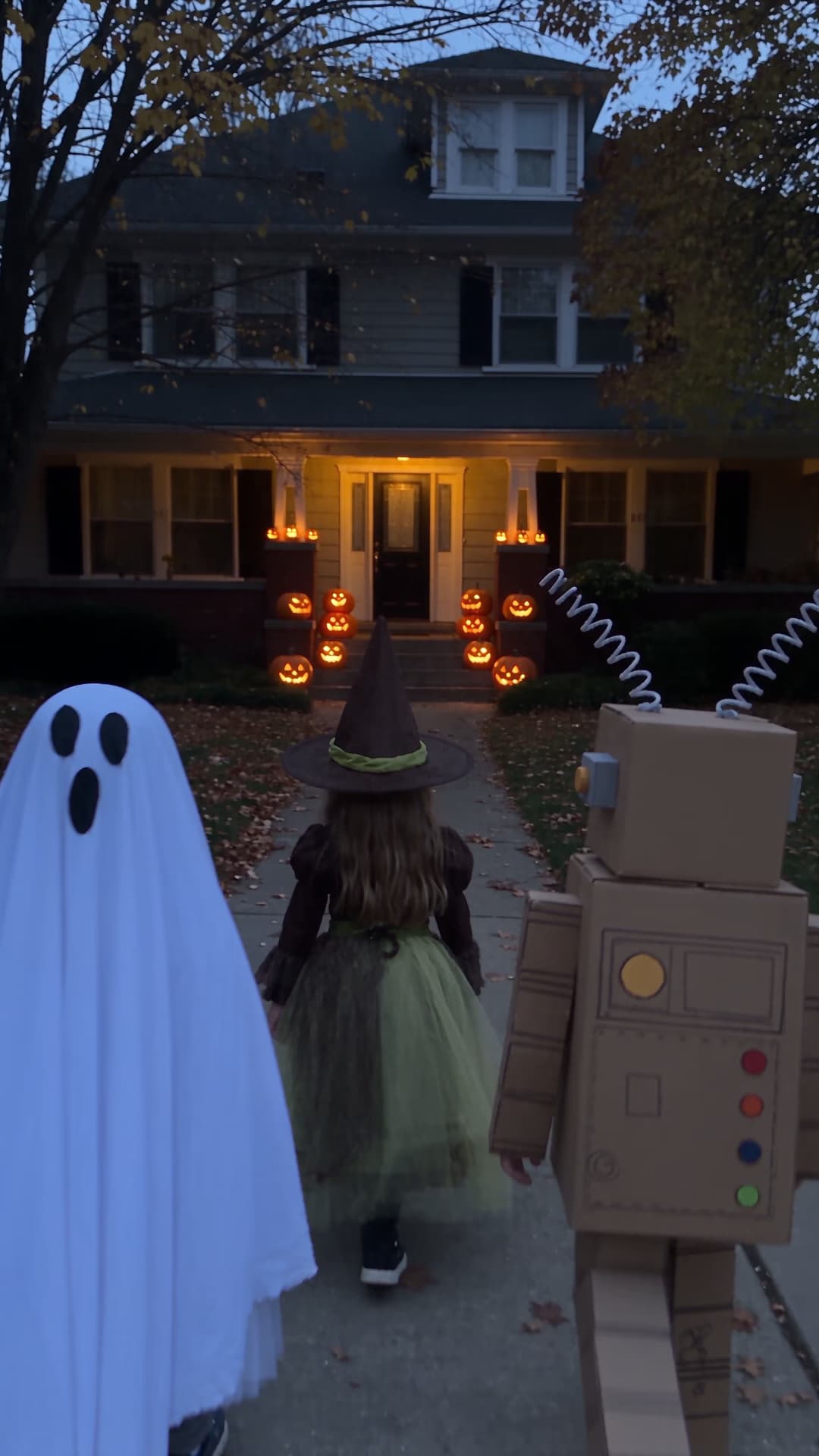 Three children in handmade ghost, garden witch, and cardboard robot costumes walking toward a pumpkin-lit porch.